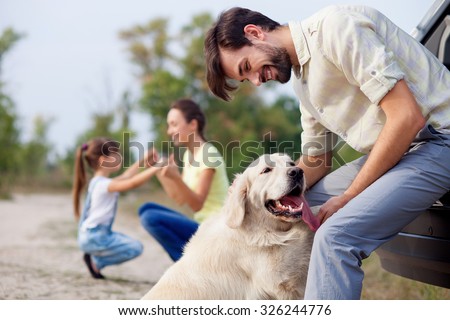 Cheerful friendly family is resting in park. The man is playing with dog and leaning on car. The mother and daughter are kneeling and holding hands. They are smiling