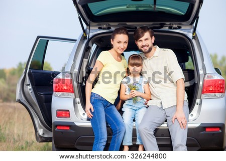 Beautiful young married couple and their daughter are sitting on boot of car outside. They are embracing and smiling. They are looking at camera happily. The small girl is holding flowers