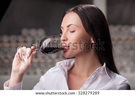 Beautiful young sommelier is drinking red wine in cellar. The woman is standing and holding wineglass. She closed eyes with pleasure