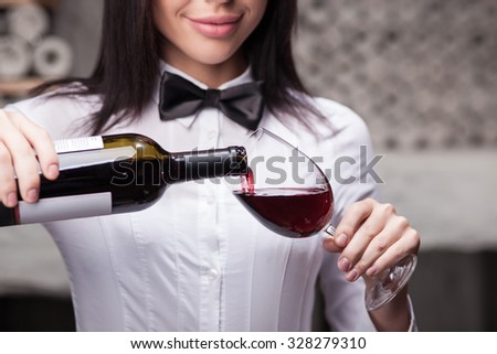 Close up of skillful young sommelier pouring red wine into glass. The woman is holding bottle and wineglass. She is standing in cellar and smiling