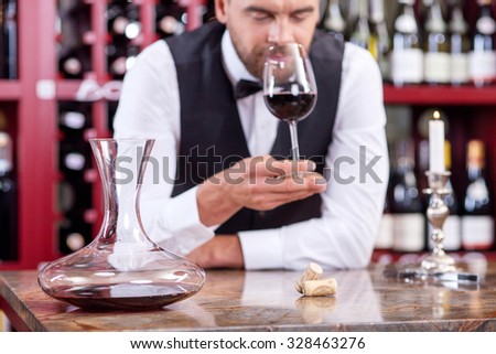 Attractive professional sommelier is smelling wine. He is holding a wineglass near his nose. His eyes are closed with pleasure. The man is standing near the table in cellar