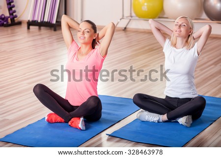 Cheerful young women are exercising at gym. They are sitting on carpets in lotus position and smiling. Their eyes are closed with relaxation