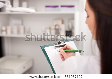 Close up of attractive young expert beautician scheduling next appointment. The woman is holding a folder of papers and writing with concentration. She is standing in her office