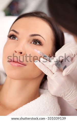 Close up of hands of expert beautician injecting botox into female face. She is standing and holding syringe. The woman is sitting and looking forward with confidence
