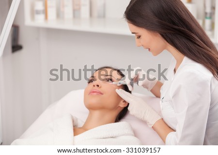 Attractive young woman is getting a collage injection in her face. She is sitting calmly at clinic. The expert beautician is filling female nasolabial wrinkles by hyaluronic acid and smiling