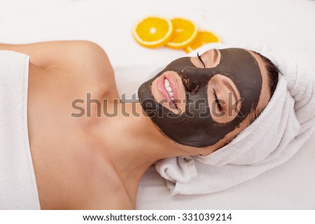 Beautiful young woman is getting facial chocolate mask at spa. She is lying with closed eyes. The girl is smiling