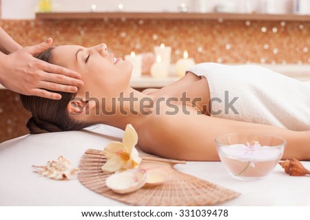 Close up of arms of masseuse massaging female head at spa. She is standing and touching hair of client. The beautiful young woman is lying and smiling