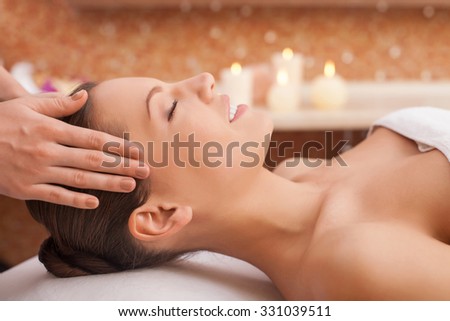 Beautiful young woman is getting head massage at spa. She is lying and smiling. Her eyes are closed with pleasure. The female hands of masseuse are massaging her carefully