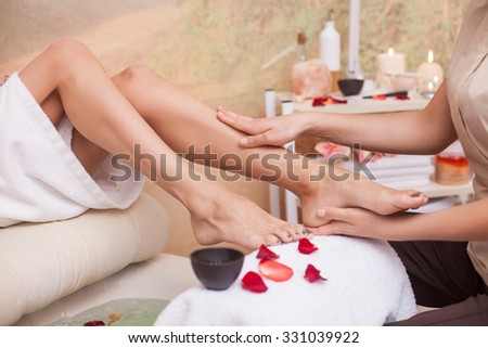 Close up of hands of masseuse massaging female legs at spa. The woman is sitting near small bath with petals