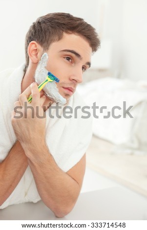 Cheerful young man is shaving in the morning. He is looking at the mirror with seriousness and holding a razor. The man is standing with a white towel on his neck