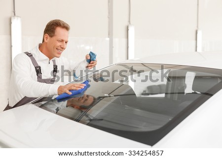 Cheerful mid aged mechanic is washing the car with a cloth. He is spraying on glass and wiping.  The man is standing in garage and smiling