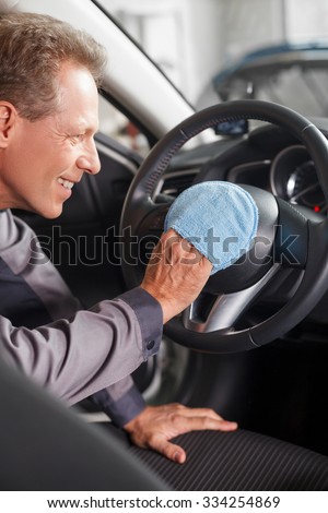 Experienced mid aged man is cleaning the steering wheel of the car. He is dusting it with cloth and smiling