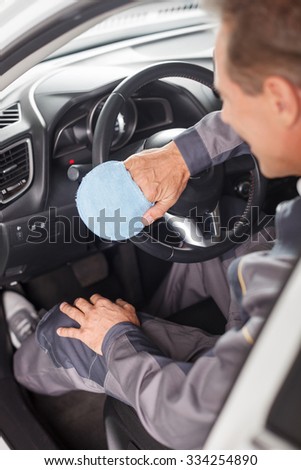 Skillful male auto service worker is cleaning the salon of the car. He is wiping dust from the steering wheel with a cloth. The man is sitting and smiling