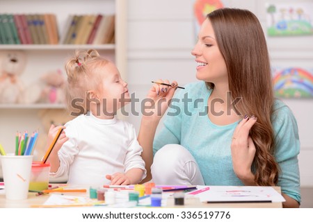 Attractive young mother is teaching her daughter painting. She is applying paint on nose of girl with fun. They are sitting at the table and laughing