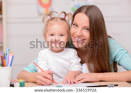 Beautiful mother is teaching her daughter to draw. They are sitting at the desk and smiling. The lady is embracing the girl