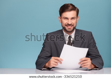 Attractive young tv newscaster is reporting news. He is sitting at the desk in a studio. The man is looking at camera and smiling. Isolated on blue background and copy space in left side
