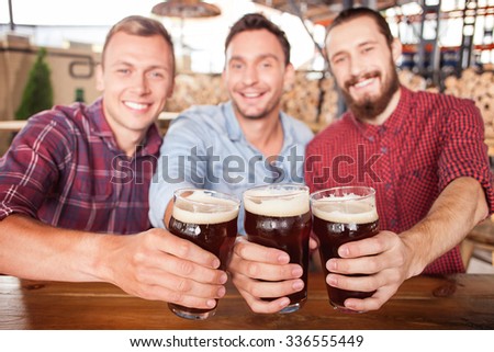Cheerful men are drinking beer in pub. They are sitting at counter and smiling. The guys are clinking glasses and looking at camera happily. Focus on three glasses of beer