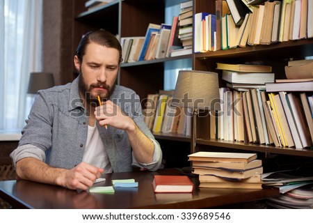 Handsome young man is studying with books at the library. He is sitting at the desk and thinking with seriousness. The student is touching a pencil to his beard