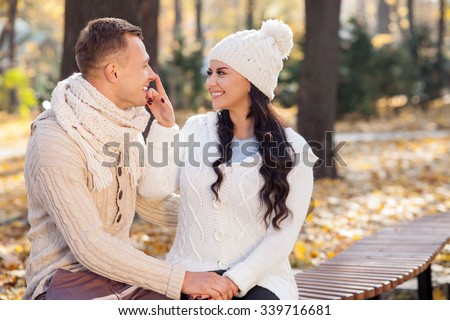Beautiful young loving couple is sitting on bench in the autumn park. They are holding hands and smiling. The woman is touching male nose playfully. They are looking at each other with love