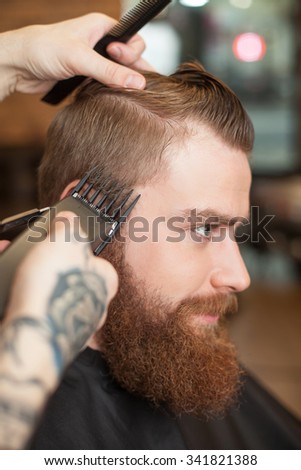 Close up of hands of experienced hairdresser holding clippers and a comb. He is cutting the hair of man carefully. The young bearded hipster is sitting with relaxation