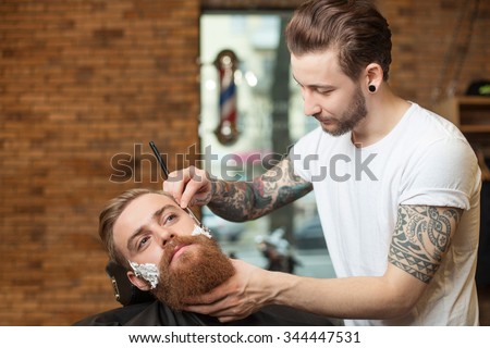 Attractive hairdresser is shaving male beard with the knife. He is standing and looking at human face with joy. The bearded man is sitting with seriousness