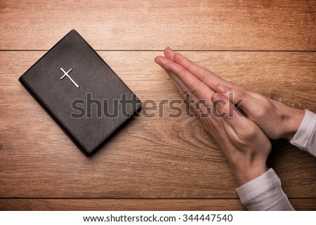 Close up of arms of young woman praying near the Bible. Her hands are clasped on the table