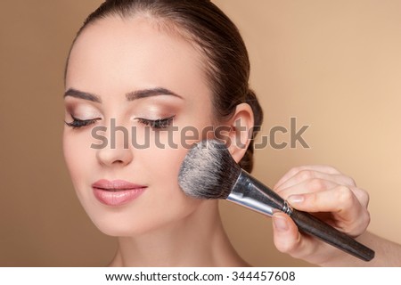 Close up of hand of professional make-up artist applying powder on female check. She is holding a brush. The woman closed eyes with pretty smile