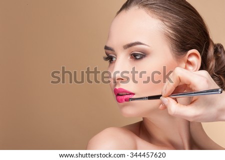 Close up of hand of visagiste applying lipstick on female lips. She is holding a brush. The young beautiful woman is looking forward with desire. Isolated and copy space in left side