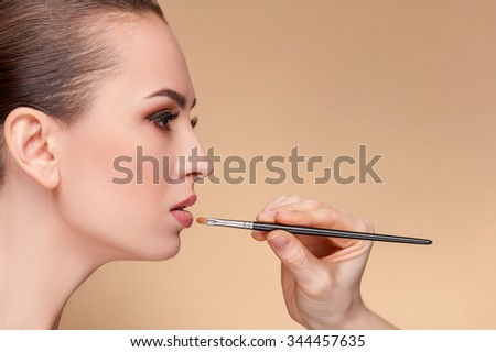 Cheerful female model is receiving treatment by make-up artist. The female hand is holding a brush and applying lipstick on her lips. The woman is looking forward with serenity. Isolated