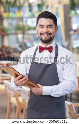 Cheerful male cafe worker is holding a menu. He is standing and smiling. The man is looking at camera happily