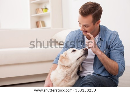 Attractive young man is sitting on flooring and embracing his dog. He is looking at him with joy and smiling