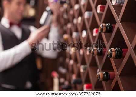 Cheerful young sommelier is choosing wine in cellar. He is smiling. Focus on bottles in shelf