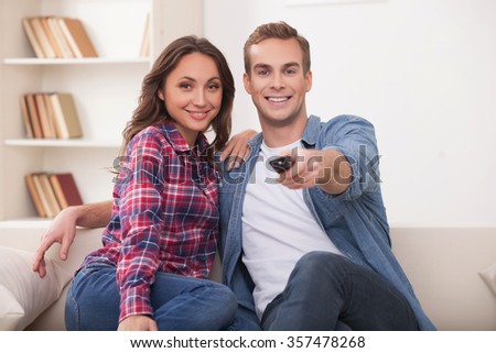 Beautiful young married couple is watching tv with joy. They are sitting on sofa and embracing. The man is holding the remote. They are smiling