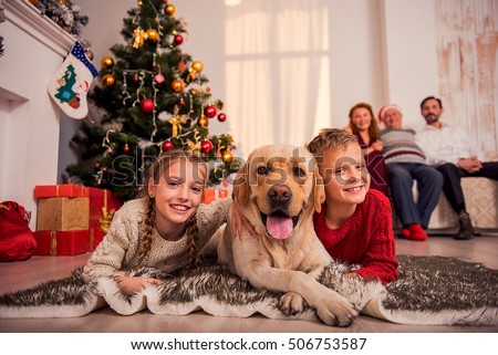 Happy children are lying on floor near Christmas tree and embracing dog. They are looking at camera and smiling. Parents are looking at them with proud