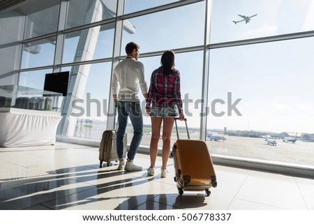 Young man and woman are looking through window at airport dreamingly. They are carrying luggage and holding hands
