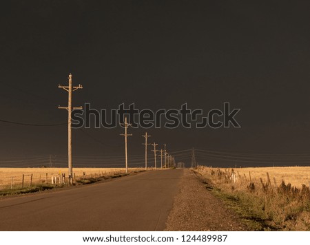 Rural road, with ominous black sky above, backlit by the evening sun.