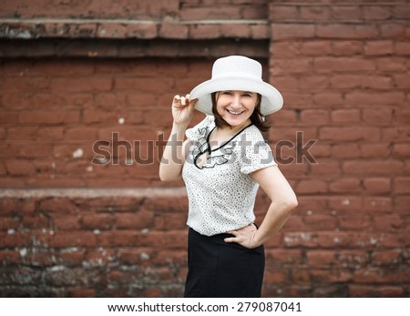 A smiling young woman in a white blouse and a hat posing against the backdrop of an old vintage brown brick wall. Female hand holds a hat.  Photo in retro style. Selective focus on model.