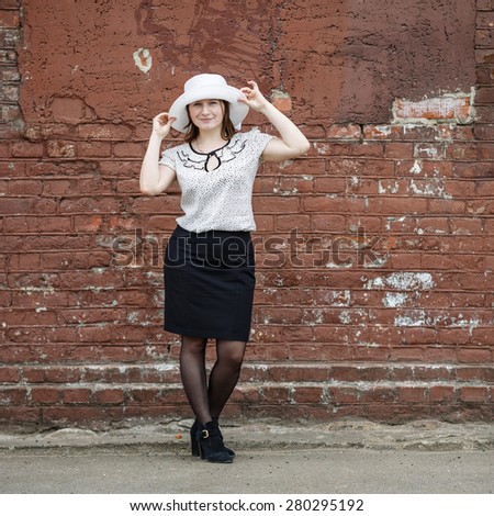 Woman in a white hat, blouse and black skirt, standing against the backdrop of an old vintage brown brick wall. Model holds the edges of the hat by hand. Portrait of a woman in a retro style.