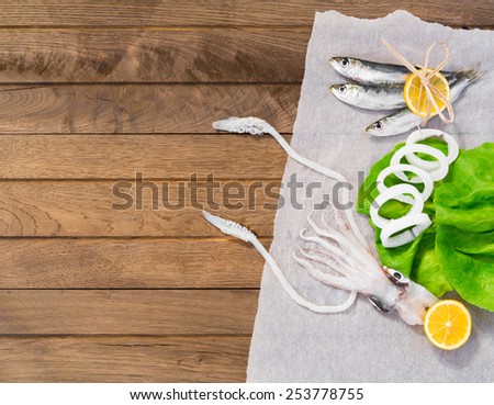 Fresh squid on wooden table, covered on half with wax paper, head with tentakles and cutted body on green lettuce, slice of lemon, three sardines, wrapped with slice of lemon. Top view.