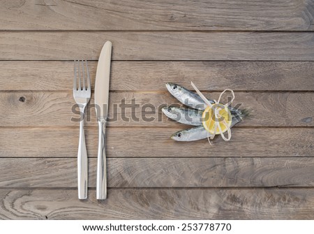 Three sardines wrapped with slice of lemon on one side and fork and knife on other side of wooden table