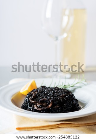 Black risotto with seafood on white plate with lemon and rosemary, bottle of white wine and glass on background