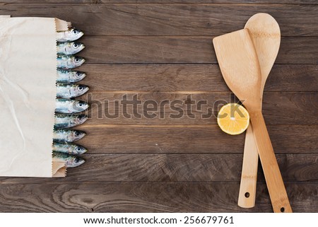 Paper bag with sardines in line on one side and kitchen utensils with lemon on other side of wooden table