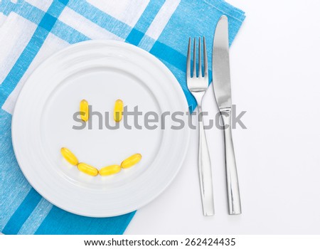 Smiley face from yellow capsules on white plate, blue and white towel, white table, fork and knife from side