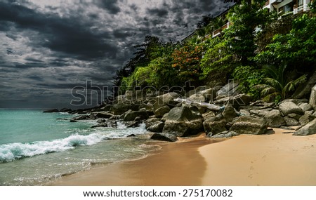 Stormy landscape with dark skyes on background and flash of sunlight on greenery, rocks and sand on foreground, house can be seen between leaves, footsteps on sand