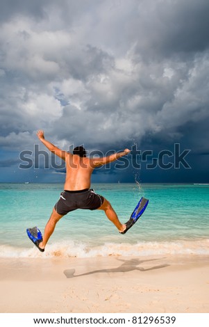 Tanned man jump in blue flippers on sand beach and ocean under gloomy sky