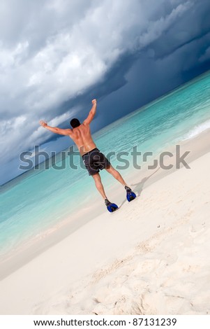 Tanned man jump in blue flippers on sand beach and ocean under gloomy sky