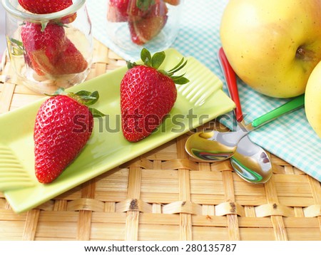 Strawberries on a green plate with colorful tea spoons on wattled surface and apples and other strawberries in the background