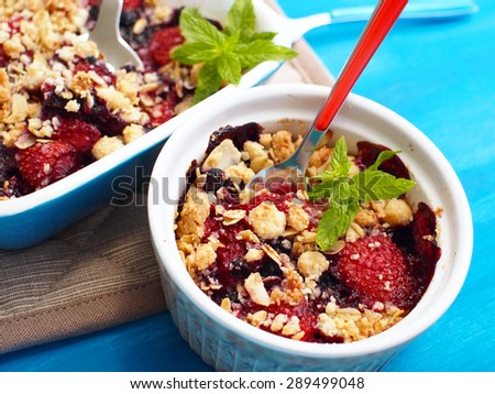 Crumble with strawberries, red currants, blueberries and blackberries with crumb made of powdered sugar, almonds, flour and oatmeal flakes in a ovenproof bowl with a spoon inside