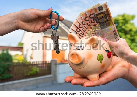 Real estate agent giving house keys to a new property owner, who is paying by euro money from piggy bank on blurred background