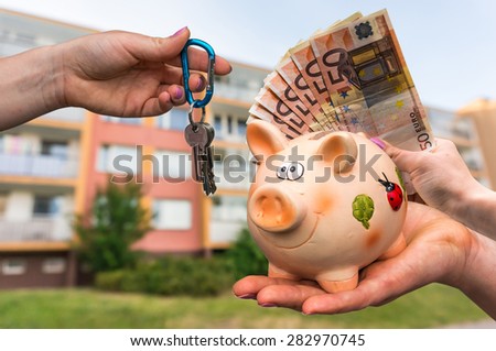 Real estate agent giving flat keys to a new property owner, who is paying by euro money banknotes from piggy bank on blurred background